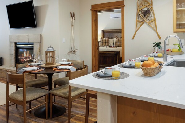 A modern kitchen with a central island, dining area with chairs, a fireplace, and a television mounted on the wall.