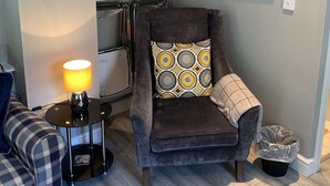 Una sala de estar acogedora con un sillón marrón, un sofá de cuadros, una mesita con una lámpara y una zona de cocina con estanterías arriba.