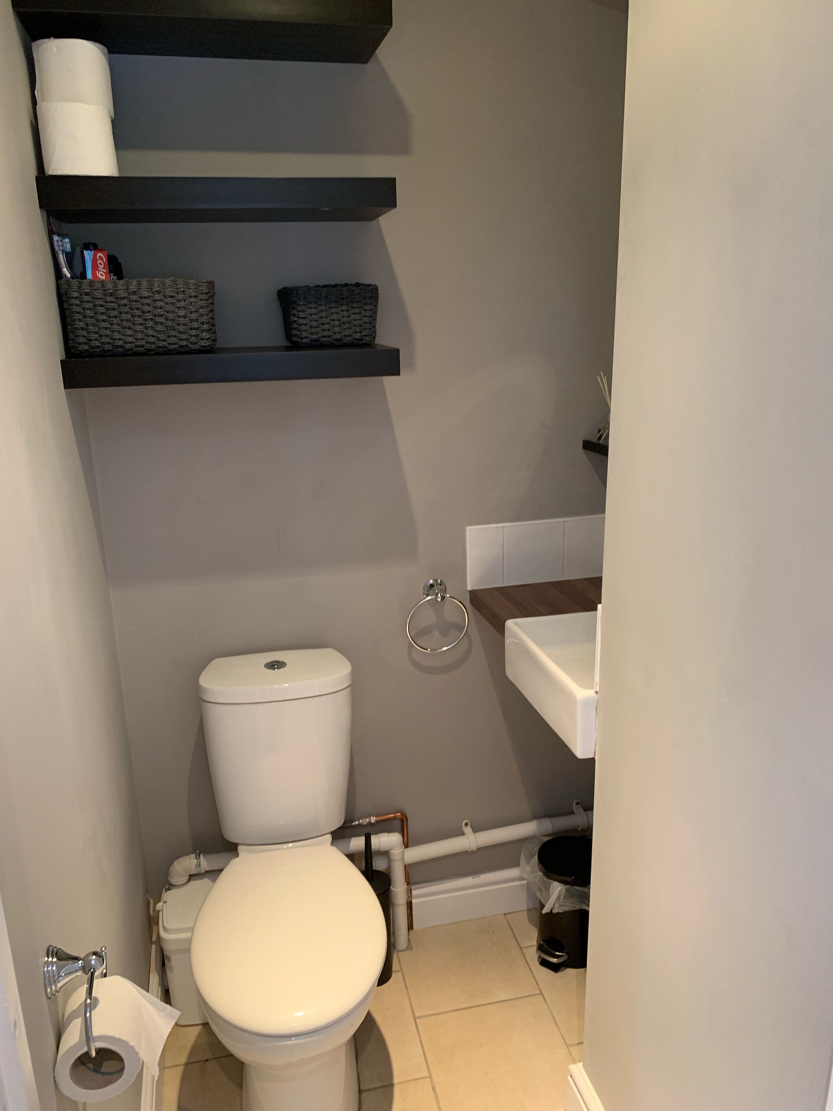 Et lille badeværelse med toilet, vask og hylder over toilettet med kurve og toiletpapir.
