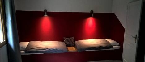 Une petite pièce, de taille modeste, avec deux lits adossés à un mur rouge, une fenêtre sur le côté gauche et une porte sur le côté droit.