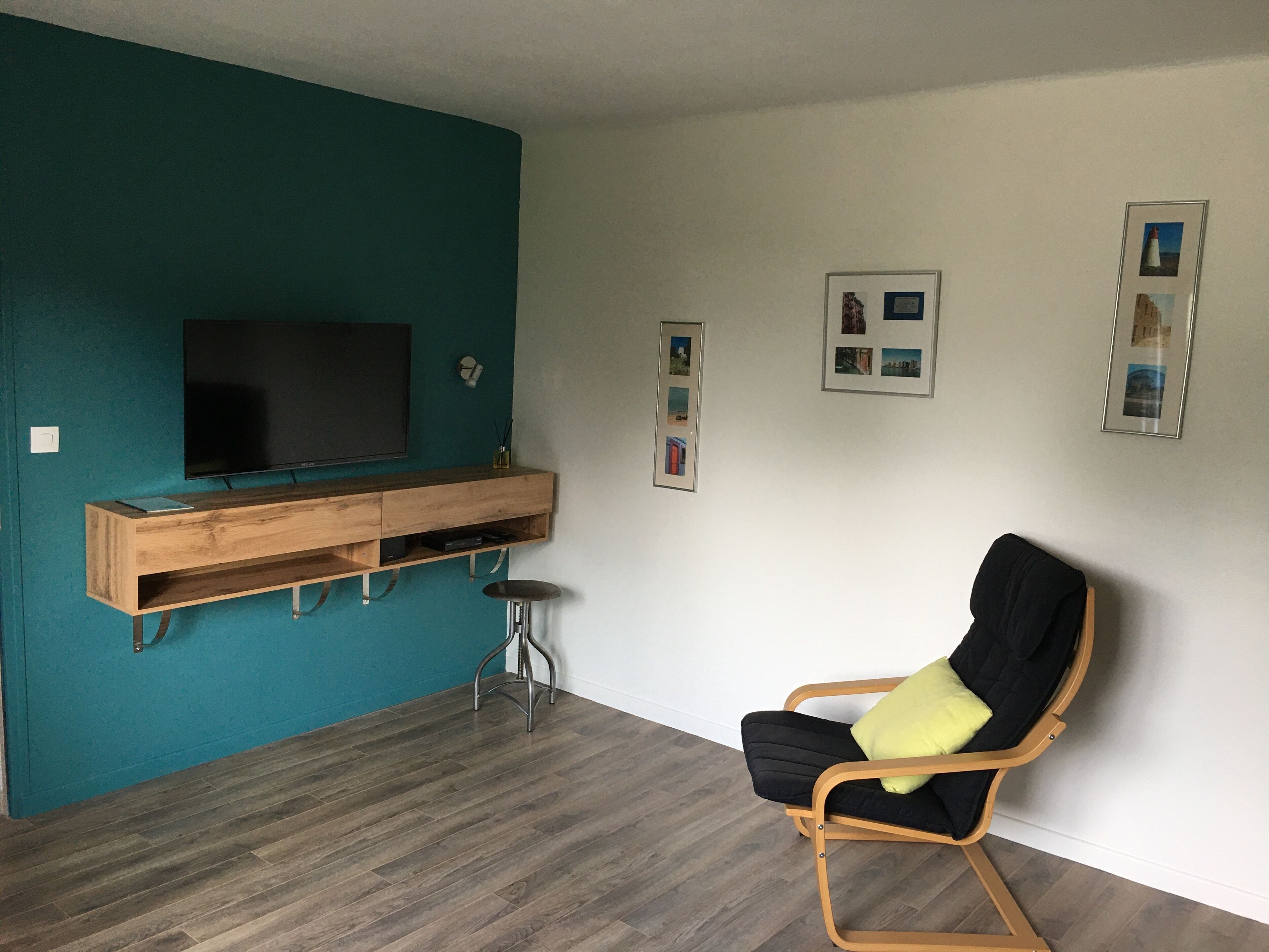 Una sala de estar con paredes color esmeralda, un mueble de madera para entretenimiento, un sofá negro con un cojín amarillo y un televisor montado en la pared.
