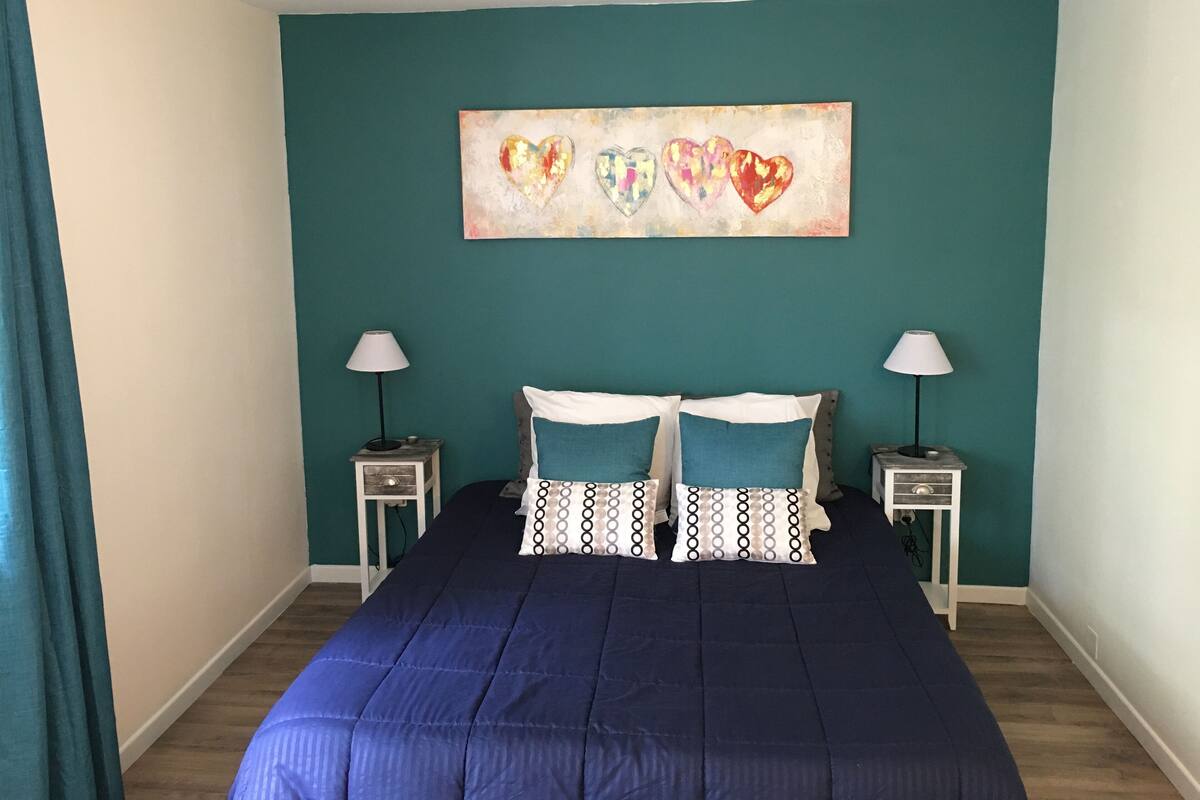 A bedroom with teal walls, a large bed with blue bedding, two bedside tables with lamps, and a heart-themed artwork above the bed.