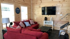 L’intérieur d’un chalet accueillant, avec un canapé rouge, un téléviseur à écran plat fixé au mur et une petite table sur laquelle se trouve une lampe.