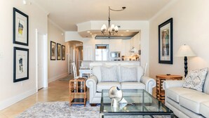 A spacious living room with a glass coffee table, two sofas, a dining area with a kitchen in the background, and framed artwork on the walls.