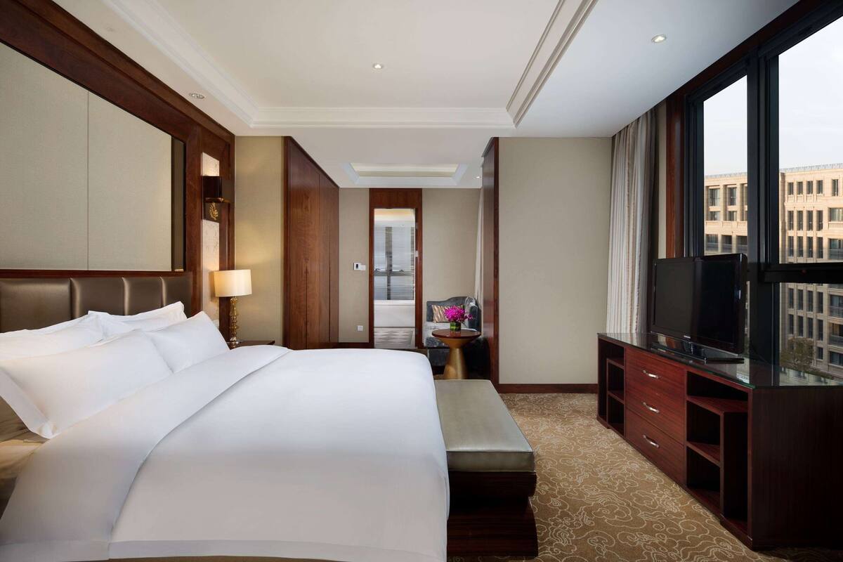 A hotel room with a large bed, a TV, a desk with a lamp, and a view of the city through large windows.