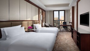 A hotel room with two beds, a desk, a chair, a TV, and a view of the city.