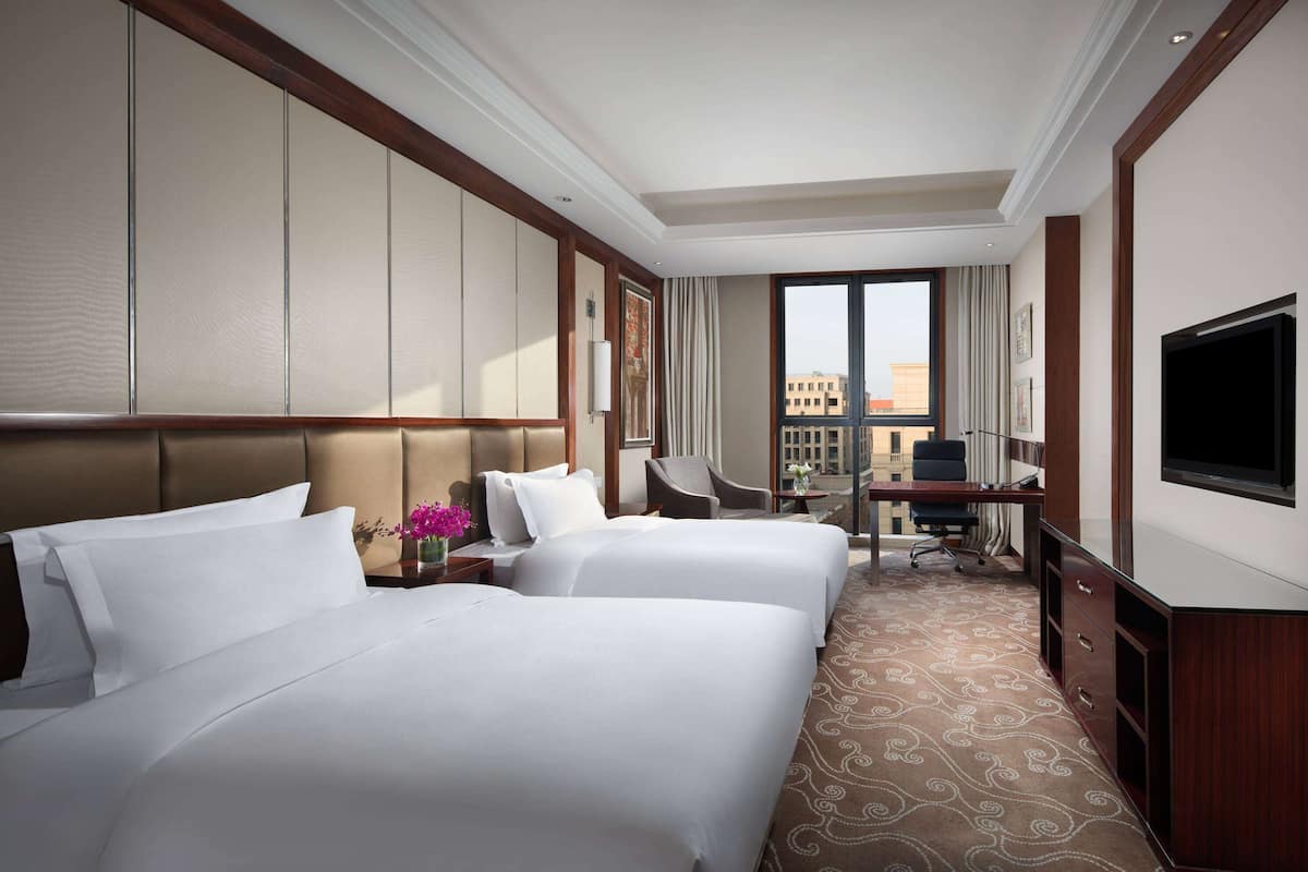 A hotel room with two beds, a desk, a chair, a TV, and a view of the city.