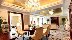 A spacious hotel lobby with a round dining table, comfortable seating, a large chandelier, and a flat-screen TV.