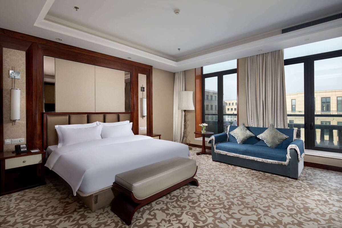 A hotel room with a large bed, a sofa, a desk with a lamp, and a view of the city through large windows.