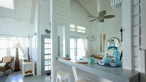 A bright, white room with a high ceiling, a ceiling fan, a bar area with stools, and a large window providing natural light.