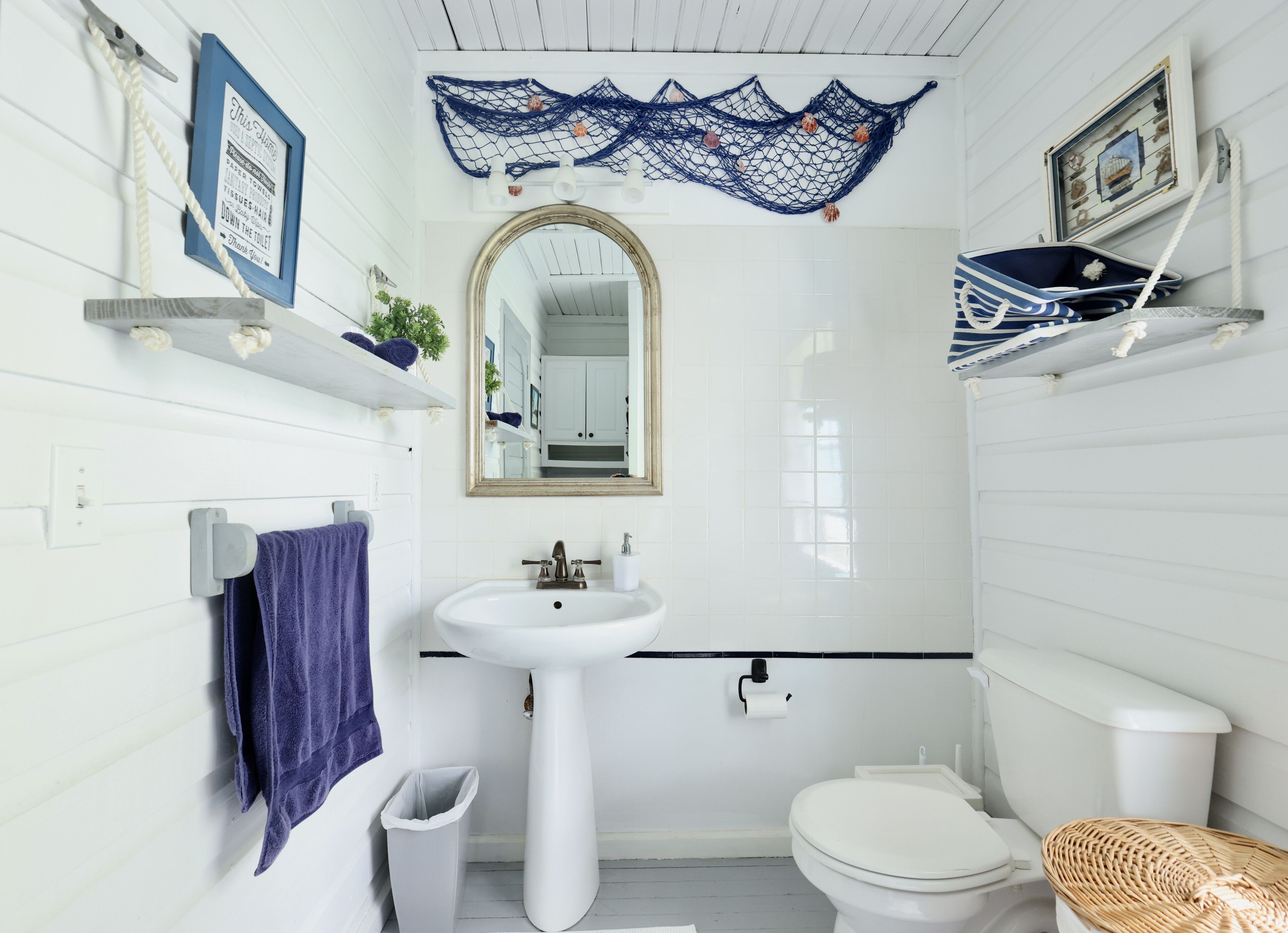 A bathroom with a white sink, toilet, and a mirror above the sink. There are shelves with towels and a trash can.