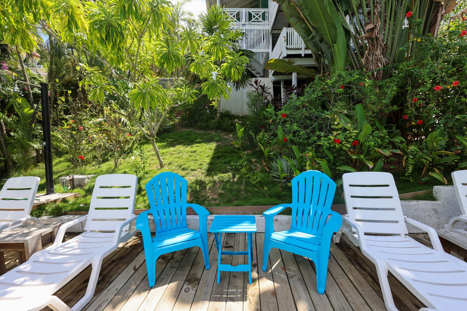 A wooden deck with white and blue lounge chairs, a small table, and a lush garden with various plants and flowers.