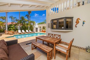 A poolside area with a wooden table and chairs, a wicker sofa, and a view of the pool and palm trees.
