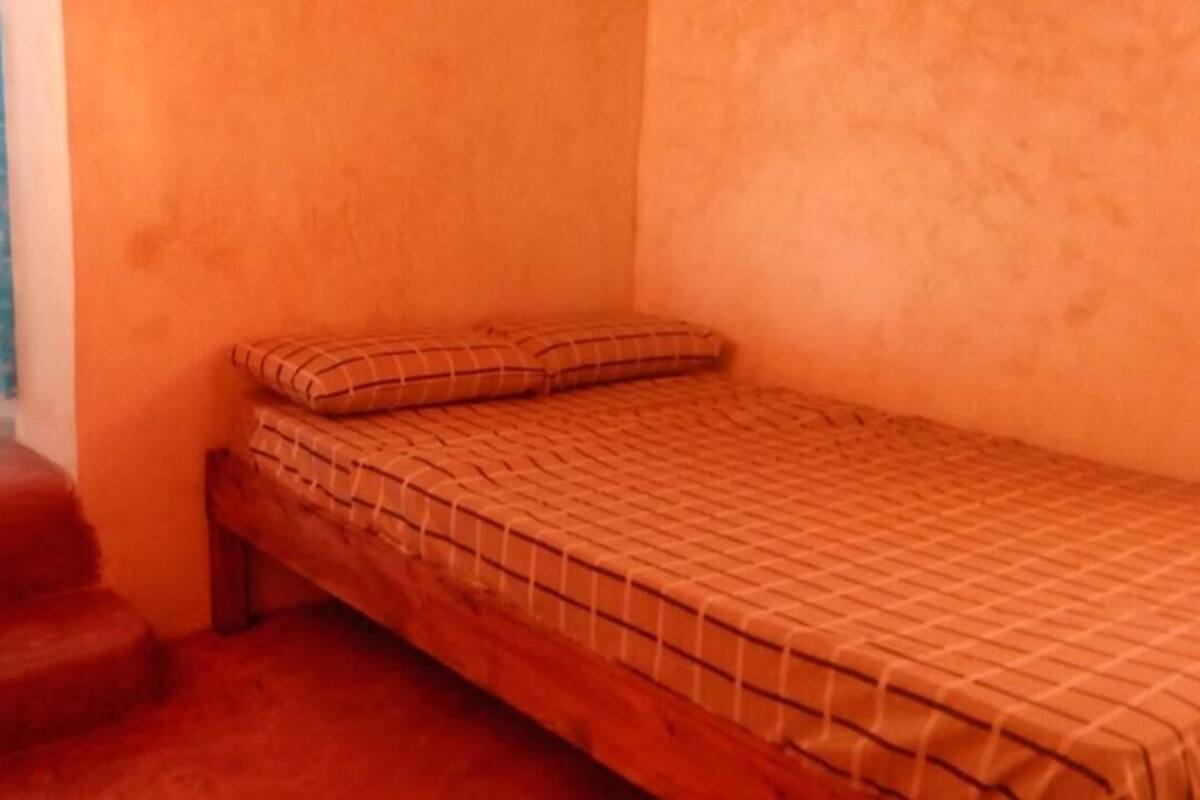 A small, single-bed room with a wooden bed frame and a checkered bedspread. There are two pillows on the bed. The room has a window with a blue curtain and a door to the left.