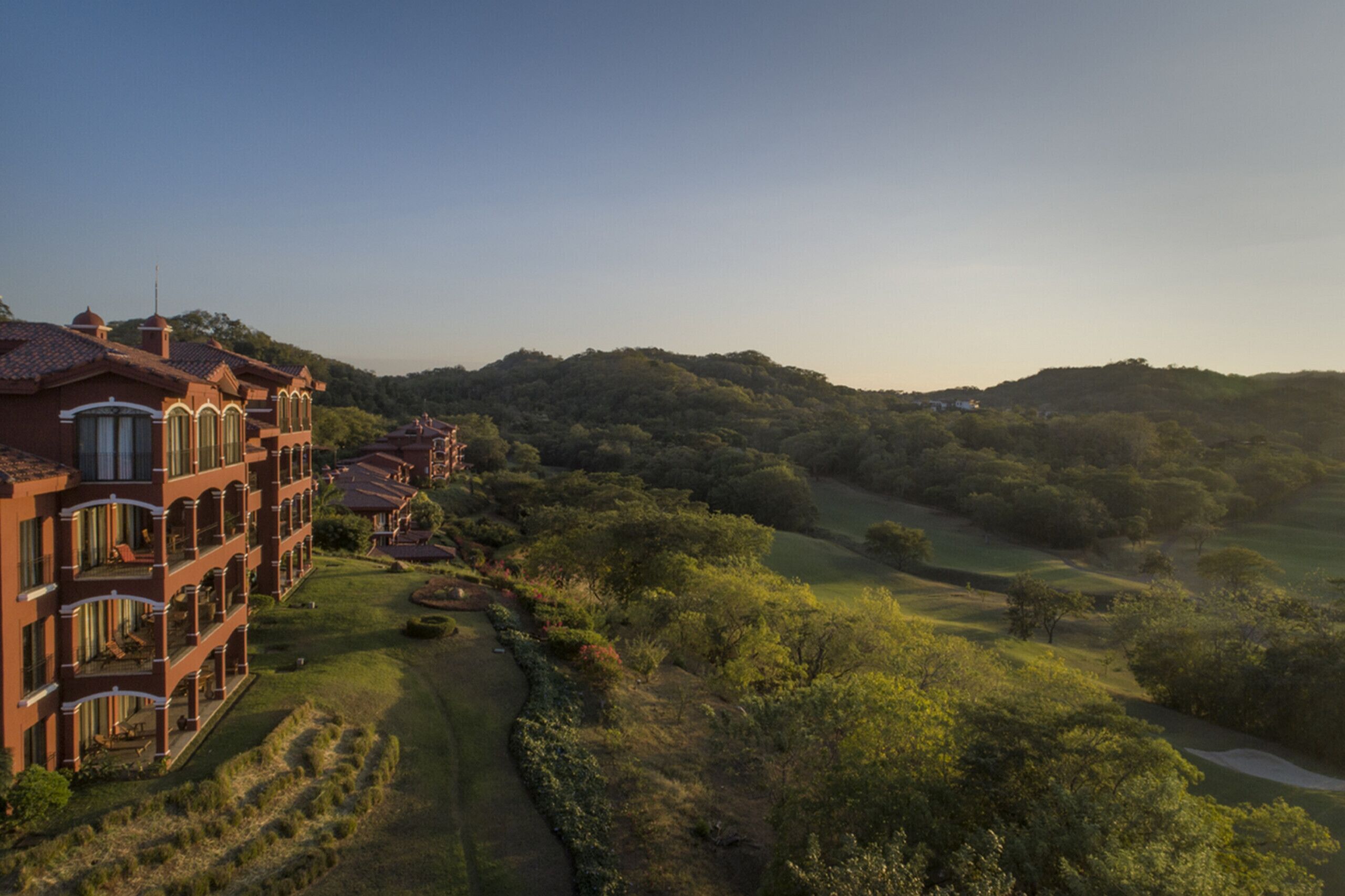 A large, multi-story hotel with a red facade and arched windows, surrounded by lush greenery and a golf course.