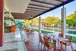 Uma área de pátio ao ar livre com mesas e cadeiras de madeira, uma piscina e um espaço de estar coberto com vista para a piscina e vegetação.