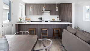 A modern kitchen with wooden cabinets, a white countertop, and pendant lights. There is a dining area with a wooden table and bar stools. - Stylish 4BD/4BA • A/C • Private Rooftop Patio • Bikes • WiFi • 2-Car Parking! (Newport Beach)