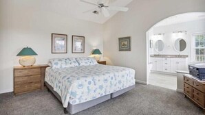 Une chambre à coucher comprenant un lit, deux tables de chevet, un ventilateur de plafond, et une salle de bain avec un lavabo et un miroir.