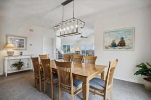 A dining area with a wooden table and chairs, a hanging light fixture, a painting on the wall, and a staircase in the background.