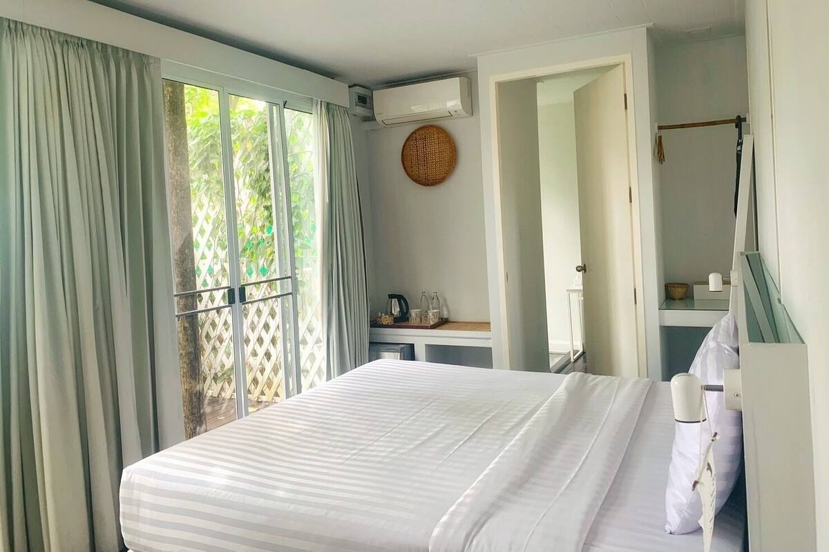 Ein Schlafzimmer mit Bett, Fenster mit Vorhängen, Klimaanlage, Nachttisch mit Wasserkocher und Spiegel.