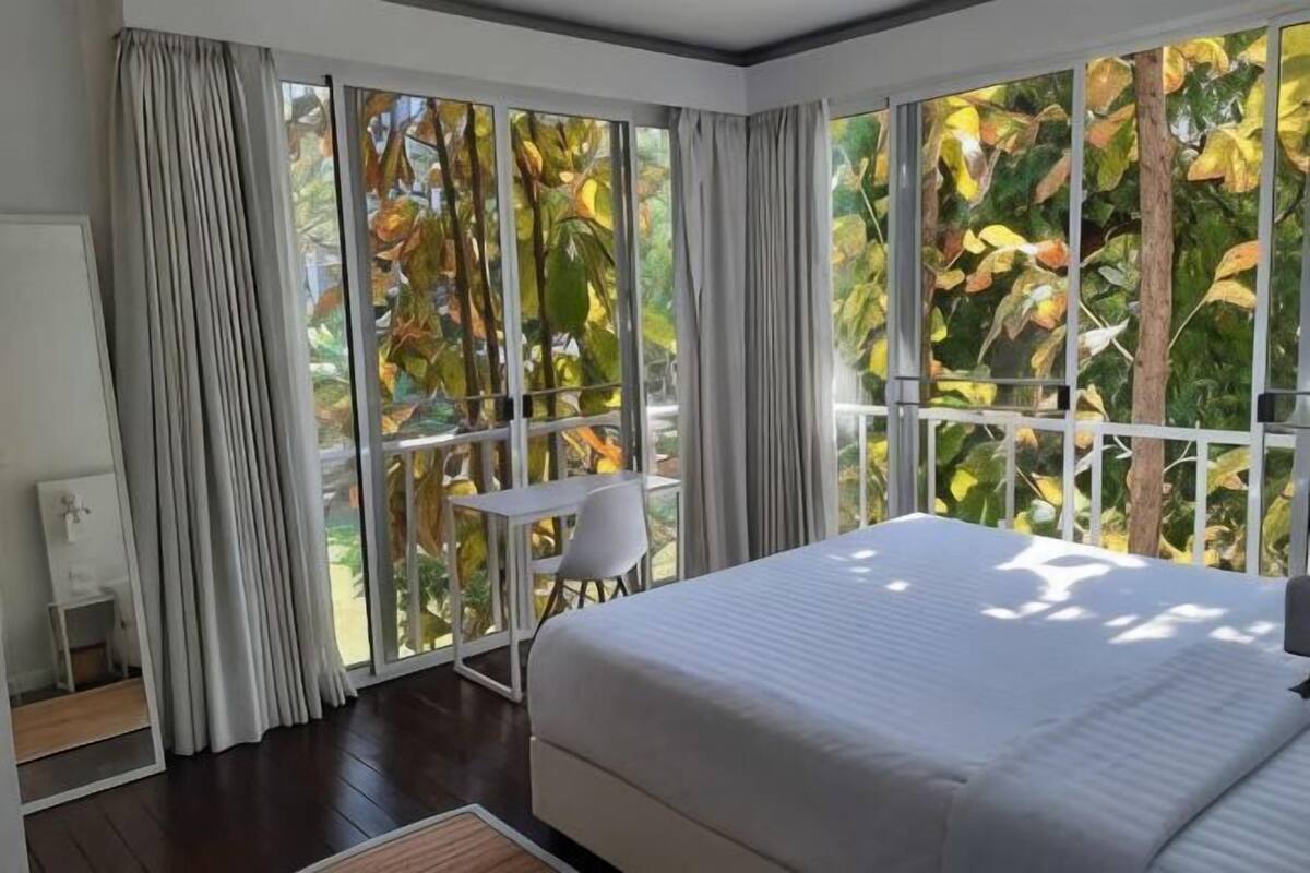 Ein Schlafzimmer mit Bett, kleinem Tisch und Stuhl neben großen Fenstern mit Blick auf Bäume.
