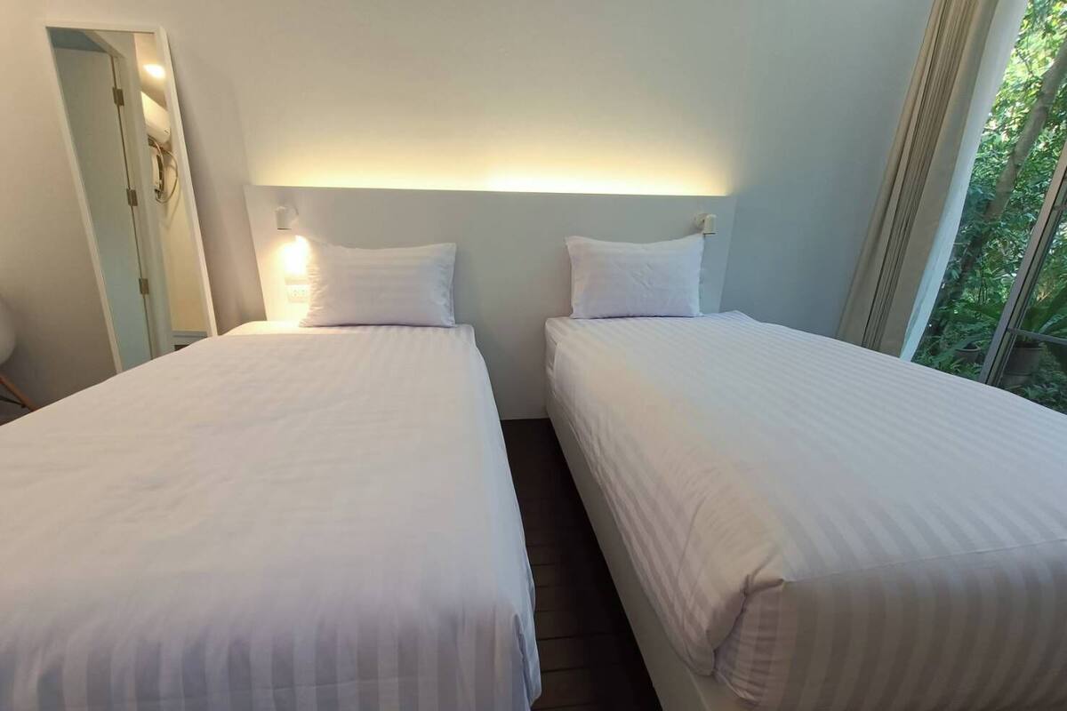 Ein Hotelzimmer mit zwei Einzelbetten, einem Nachttisch mit Lampe und einem Fenster mit Blick auf Grünflächen.