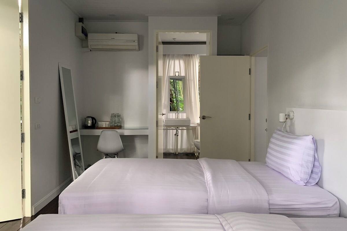 Ein Hotelzimmer mit Bett, Schreibtisch mit Stuhl, Spiegel, Klimaanlage, Fenster mit Vorhängen und einem durchgehenden Türspalt sichtbaren Badezimmer.