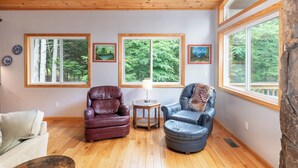 A cozy room with wooden flooring, two leather chairs, a small table with a lamp, and large windows offering forest views.