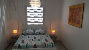 A bedroom with a bed, two bedside lamps, a window with blinds, a wall-mounted painting, and a ceiling light fixture. - Villa entre Ciel et Mer (Sainte-Anne)