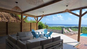An outdoor patio with a wooden deck, a seating area with a table and chairs, and a view of the ocean. - Villa entre Ciel et Mer (Sainte-Anne)
