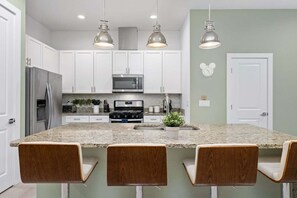 A modern kitchen with white cabinets, a granite countertop, a stainless steel refrigerator, a microwave, a stove, and three bar stools.
