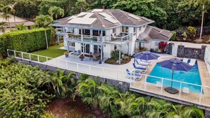 Une maison à deux étages avec une piscine, entourée d’une végétation luxuriante et sous un ciel dégagé.