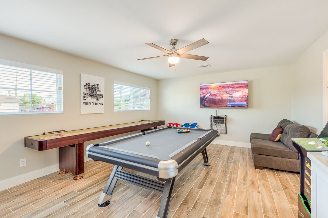 A pool table with a ceiling fan, a sofa, and a television in a room with wooden flooring and windows.