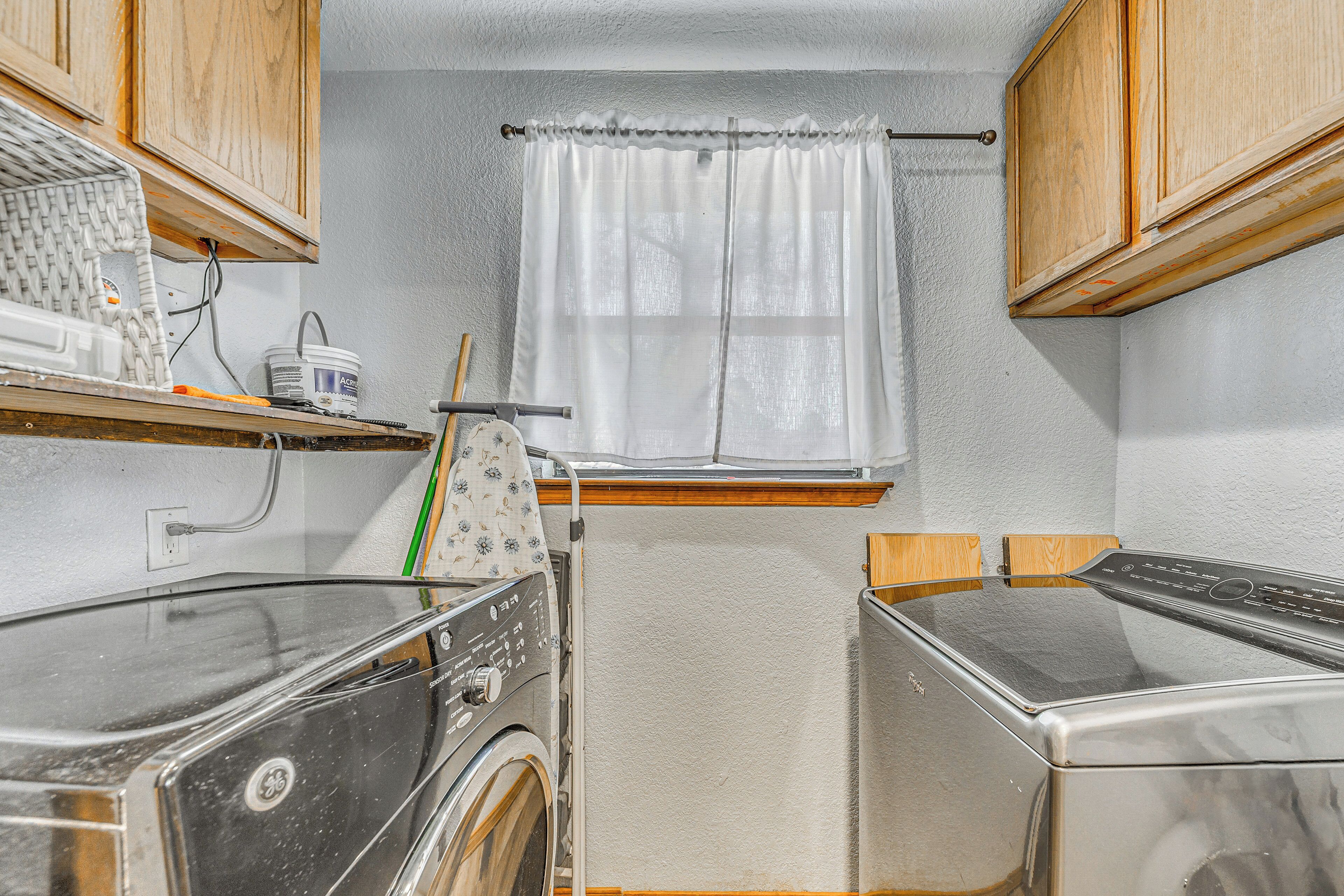 A compact laundry room with two front-loading washers, wooden cabinets, a window with a white curtain, and a small shelf with a washing machine and other items.