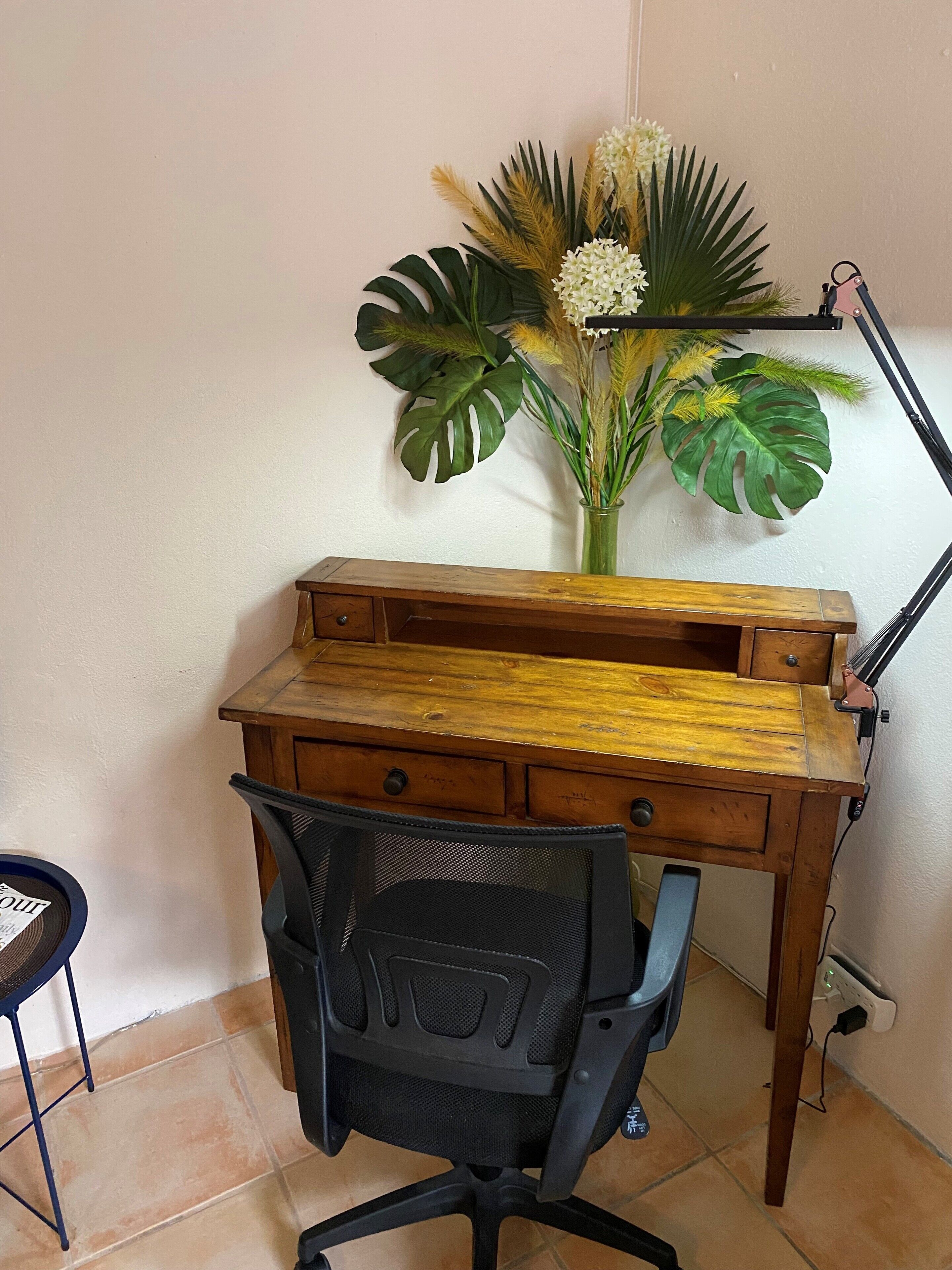 A wooden desk with a mesh chair, a vase with flowers, and a lamp.