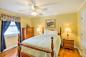 Quarto com uma cama de dossel de madeira, ventoinha no teto, mesa de cabeceira com luminária, cômoda e janela com cortinas.