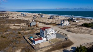 Vista aérea de una zona costera con varios edificios, incluyendo una estructura blanca destacada con techo rojo, ubicada cerca de una playa de arena y el océano.