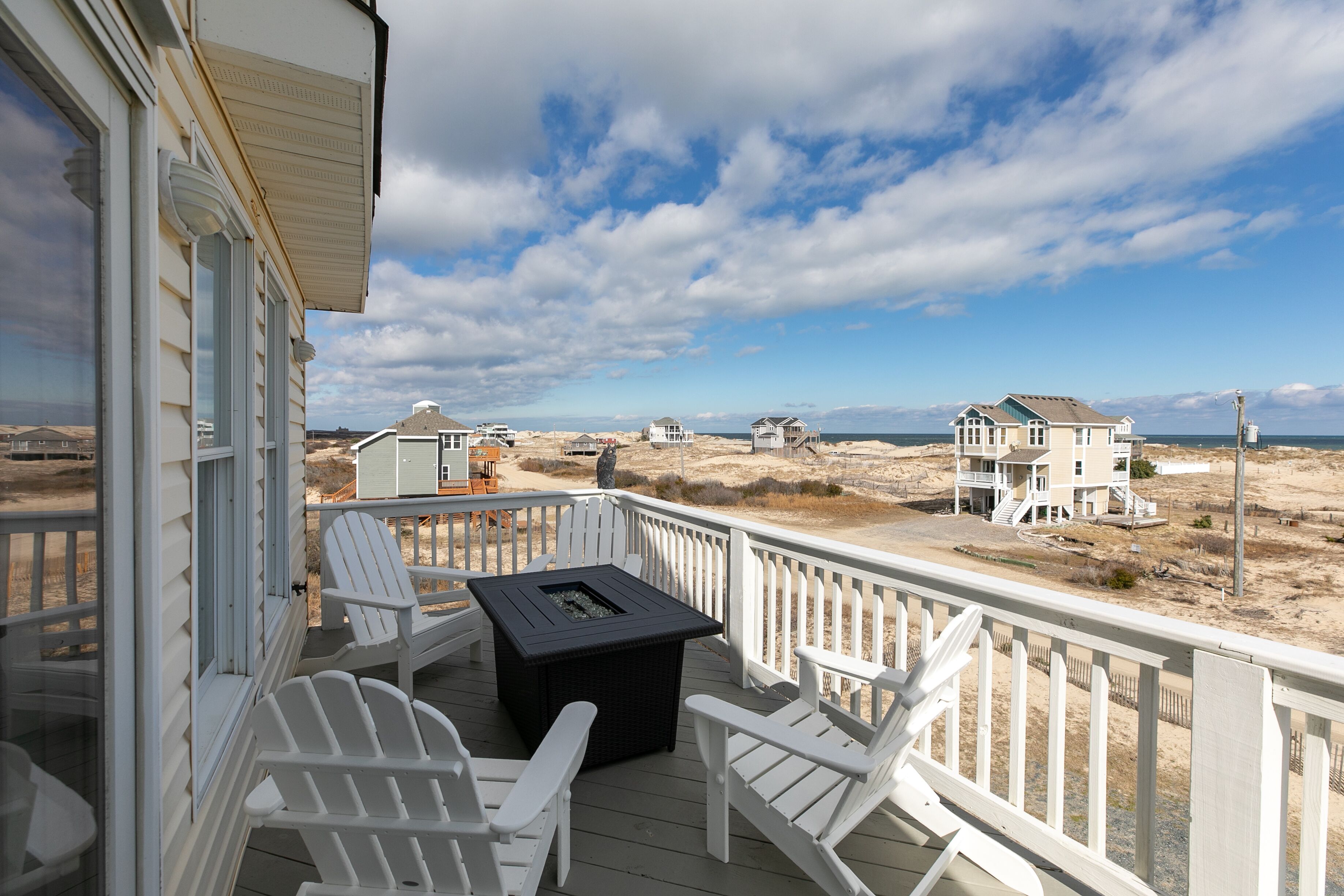 A deck with white Adirondack chairs and a black table, overlooking a beach with houses and dunes.