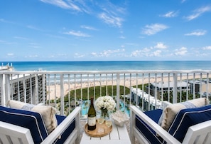A balcony with a view of the beach, a bottle of wine, and a vase of flowers.