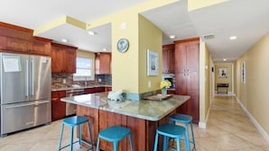 A modern kitchen with wooden cabinets, a stainless steel refrigerator, a marble island with bar stools, and a yellow accent wall. - Direct Beachfront Unit - Shores of Madeira #505 (Madeira Beach)