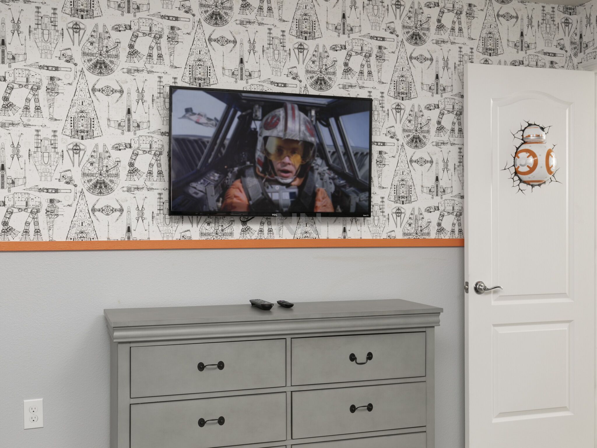 A room with a wall-mounted TV displaying a scene, a grey dresser with drawers, and a door with a sticker.