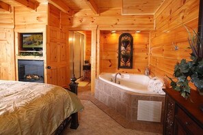 Ein Schlafzimmer in einer Blockhütte mit einem großen Bett, einem Kamin, einem Fernseher und einem Badezimmer mit Badewanne und Waschbecken.