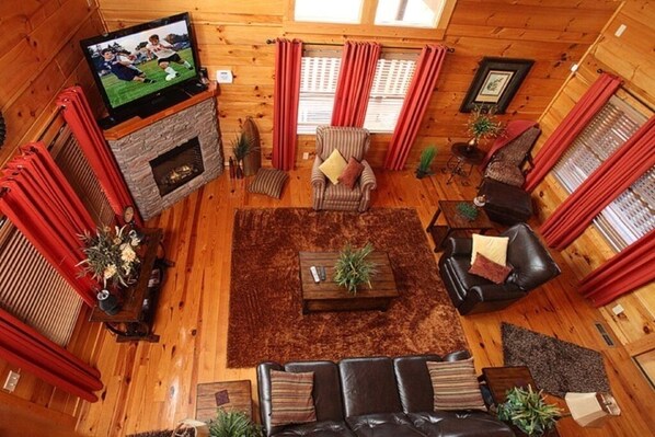 A cozy log cabin interior with a fireplace, leather sofas, a television, and red curtains.