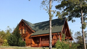 A log cabin with a prominent front porch and a steeply pitched roof, surrounded by trees and a rocky landscape. - Beautiful, clean, comfortable and great views!!  Romantic and family friendly (Sevierville)