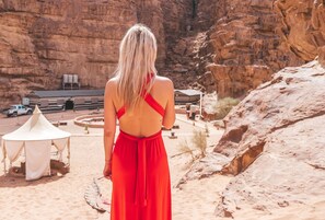 A woman in a red dress standing in a desert area with rocky cliffs and a small building in the background. - Aladdin Camp (Wadi Rum)
