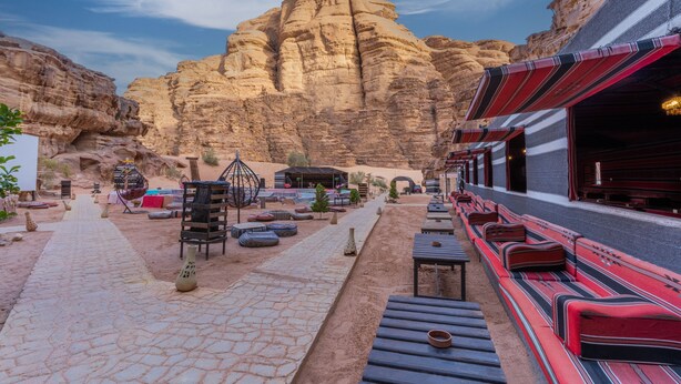 Un resort en el desierto con mesas al aire libre, una formación rocosa de fondo y un edificio con toldos a rayas.
