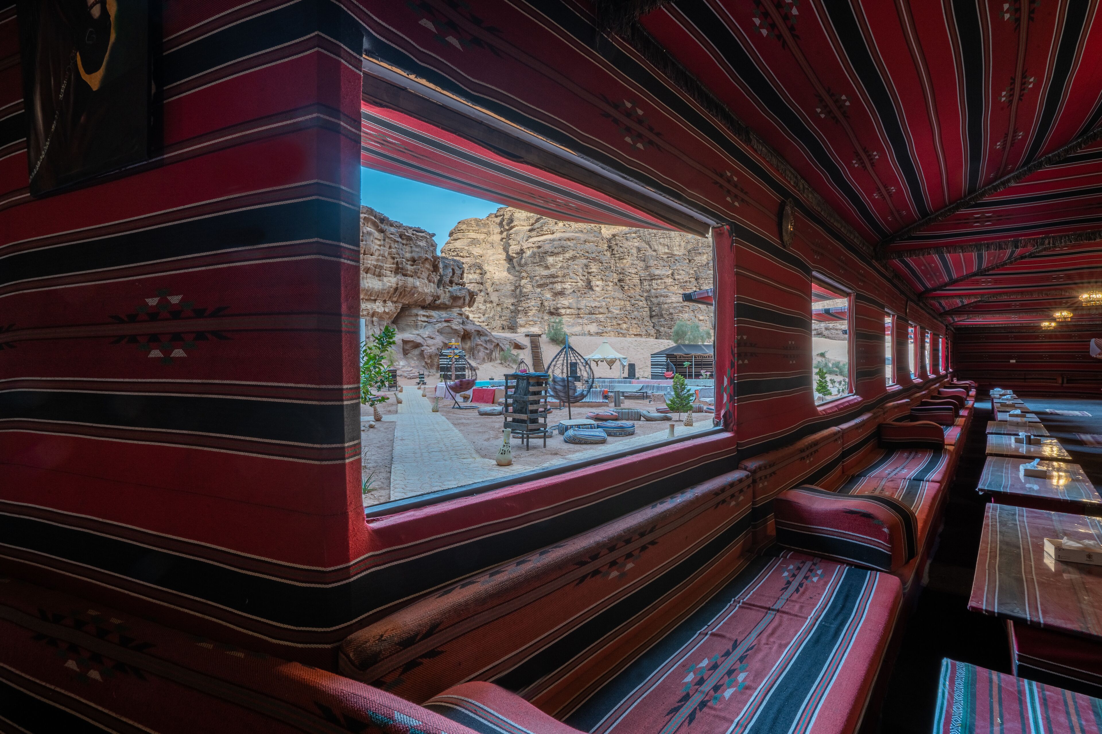 Un vagón de tren con paredes a rayas rojas, bancos de madera y vista al terreno rocoso.