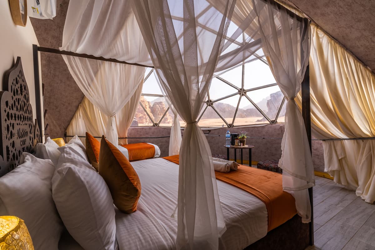 A bed with white and orange bedding, a canopy, and a view of a desert landscape.