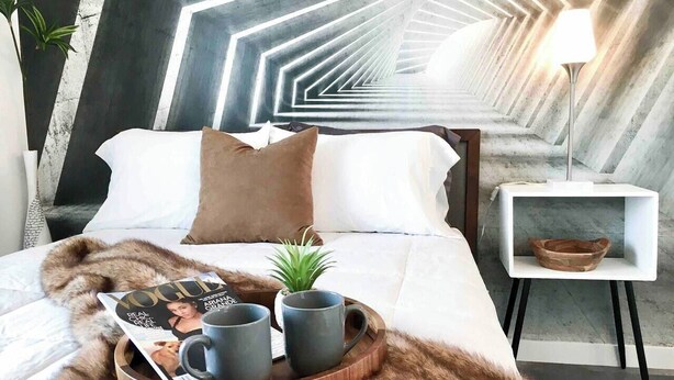 Un dormitorio moderno con techo de diseño geométrico, una mesita de madera con un cuenco, y una cama con manta de piel y una revista.