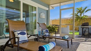 A covered patio area with wicker lounge chairs, a small table, and a view of a wooden fence and greenery.